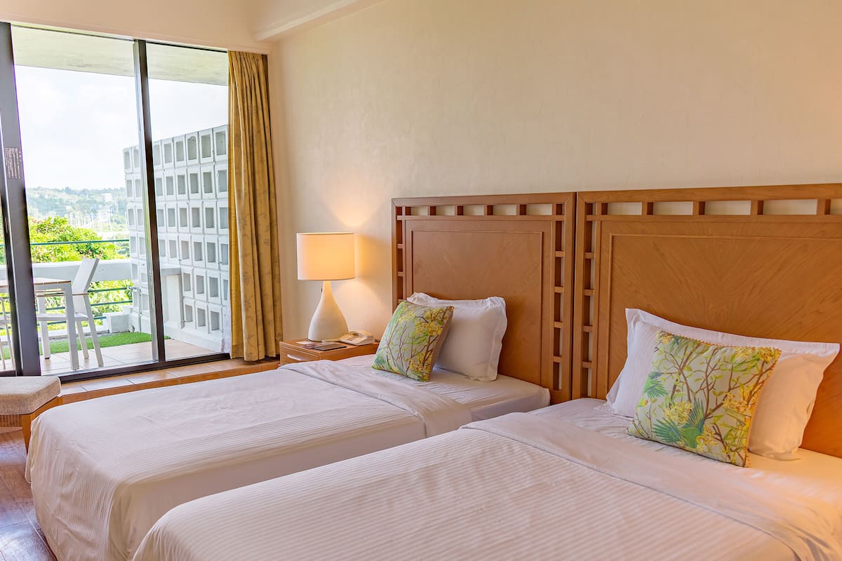 A hotel room with two beds, a nightstand with a lamp, and a view of a building through the window.