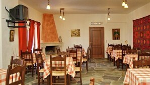 A dining area with checkered tablecloths, wooden chairs, stone flooring, a fireplace, and hanging lights. - Marina Hotel (Anogia)
