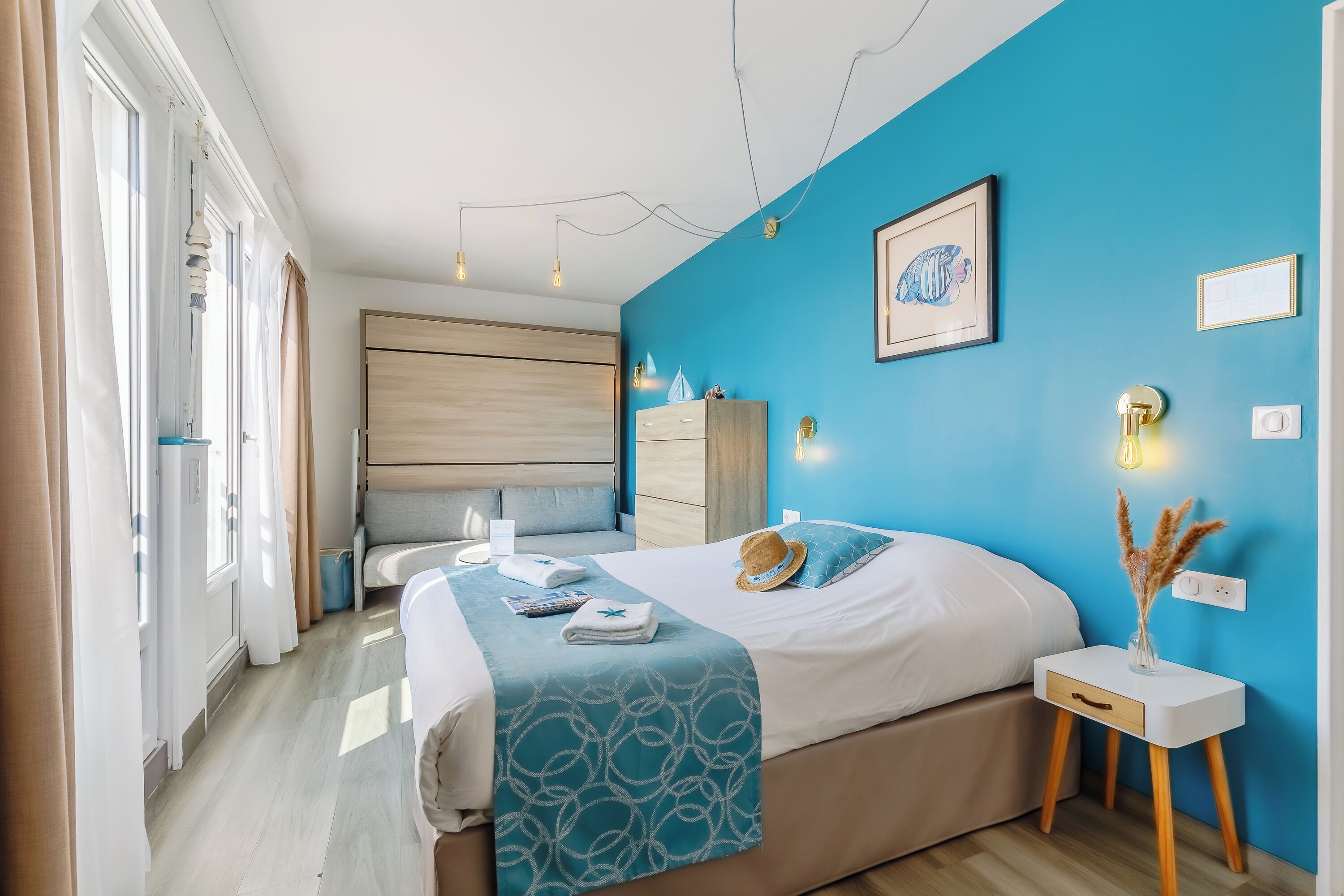 Un dormitorio moderno con una pared azul de acento, una cama con colcha de diseño azul y blanco, una mesita de noche con un jarrón y una mesita de cabecera con una lámpara.