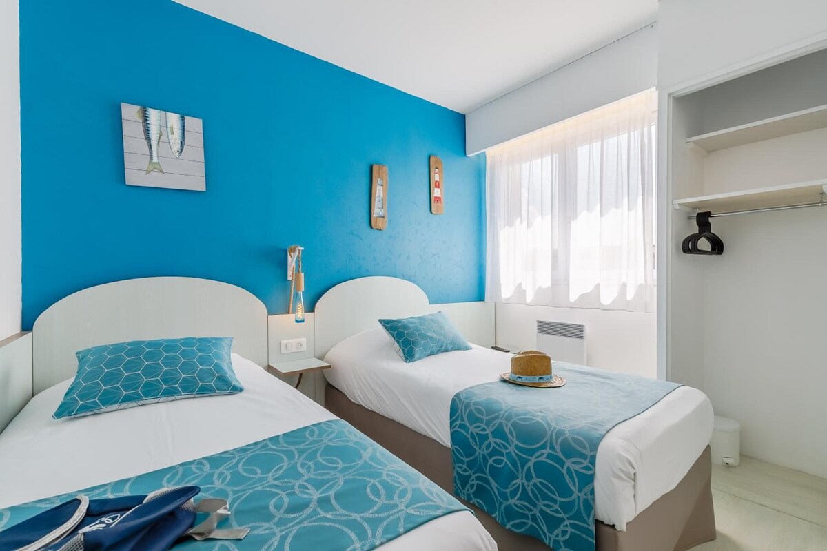 Habitación de hotel con dos camas, una pared azul, ventana con cortinas y un estante pequeño con un sombrero.