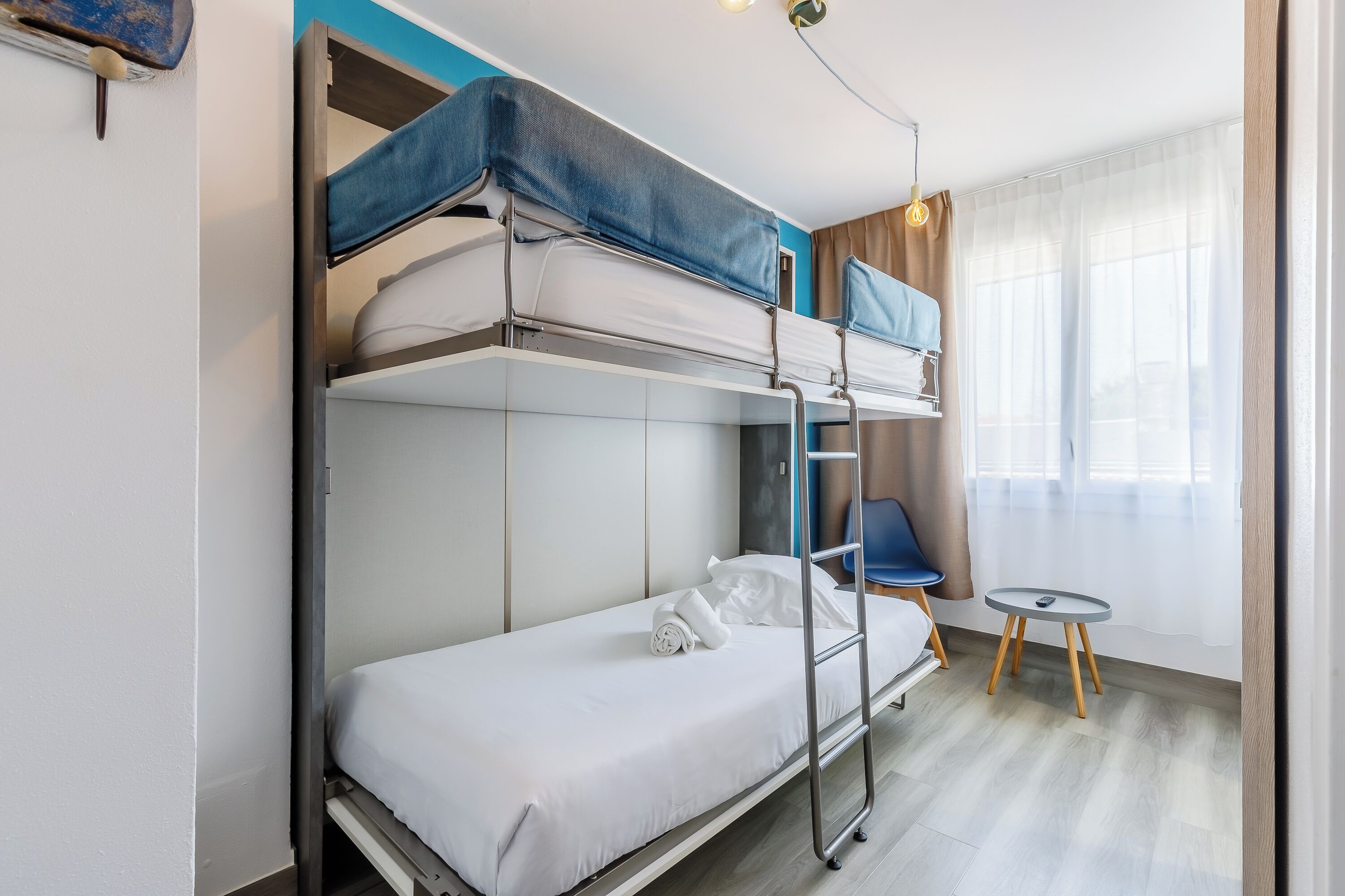 A bunk bed room with a small table and chair, a window with curtains, and a light fixture.
