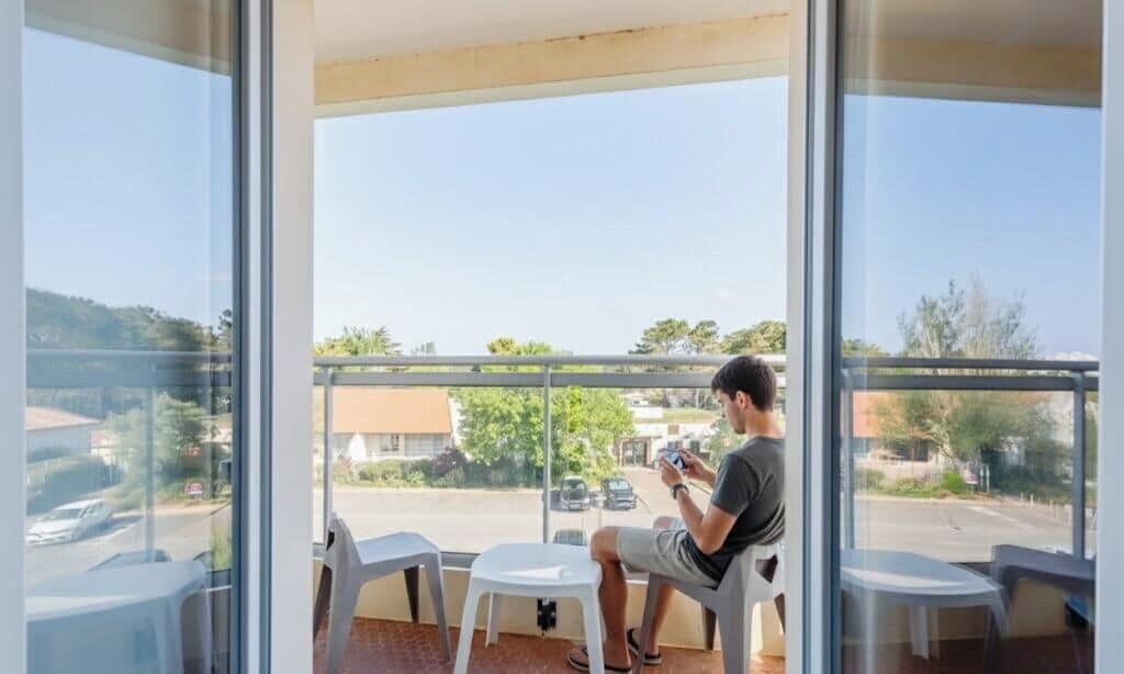 A balcony with a view of a street and buildings, a person sitting on a chair, and a table with chairs.