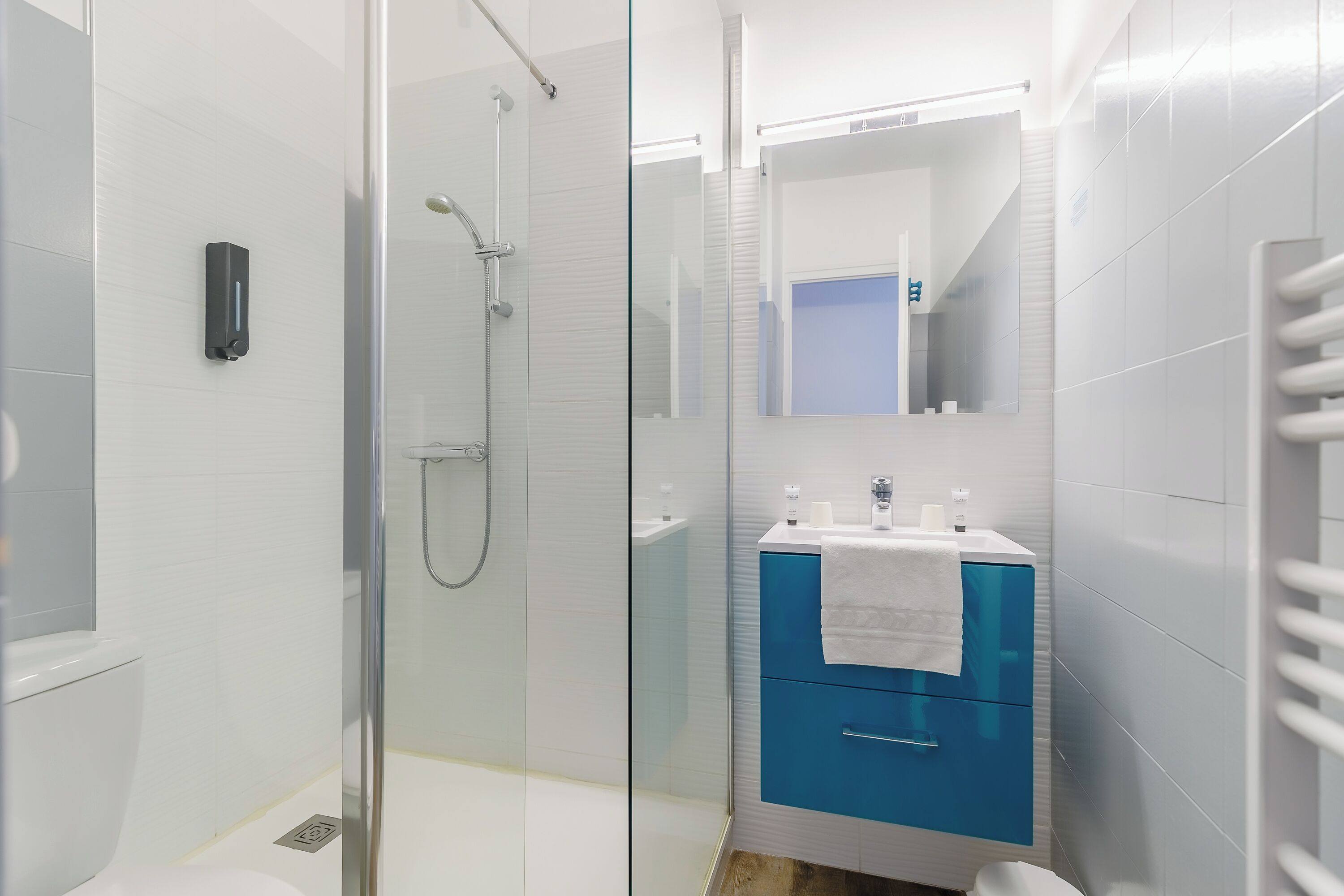 A modern bathroom with a glass-enclosed shower, a white toilet, a blue vanity with a mirror, and a towel hanging on the wall.