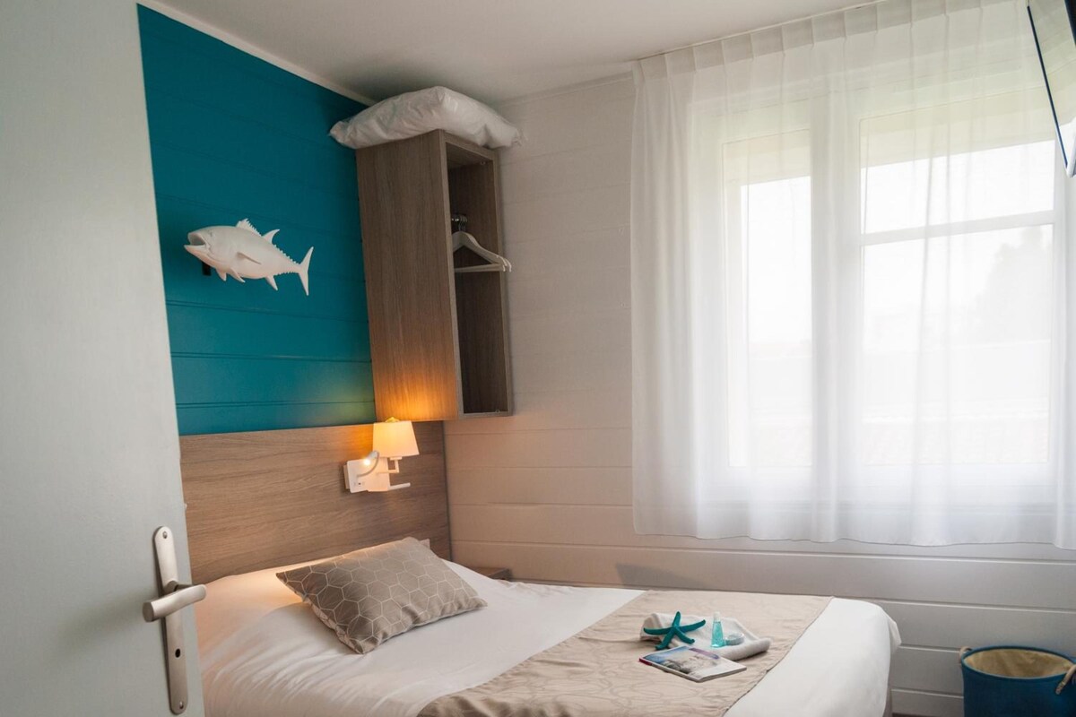 Habitación de hotel con cama, repisa en la pared, ventana con cortinas y un cubo azul pequeño.