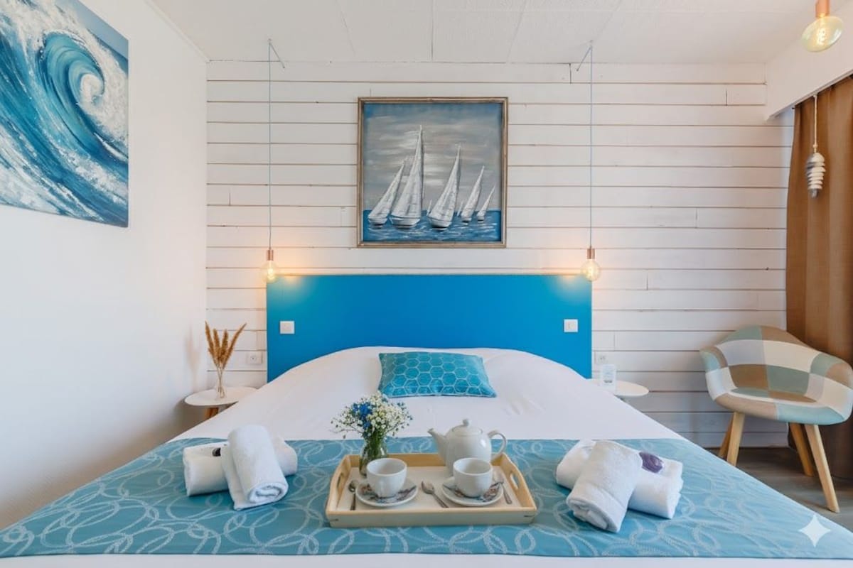 Habitación de hotel con cama, bandeja con juego de té, pintura de veleros y una silla.