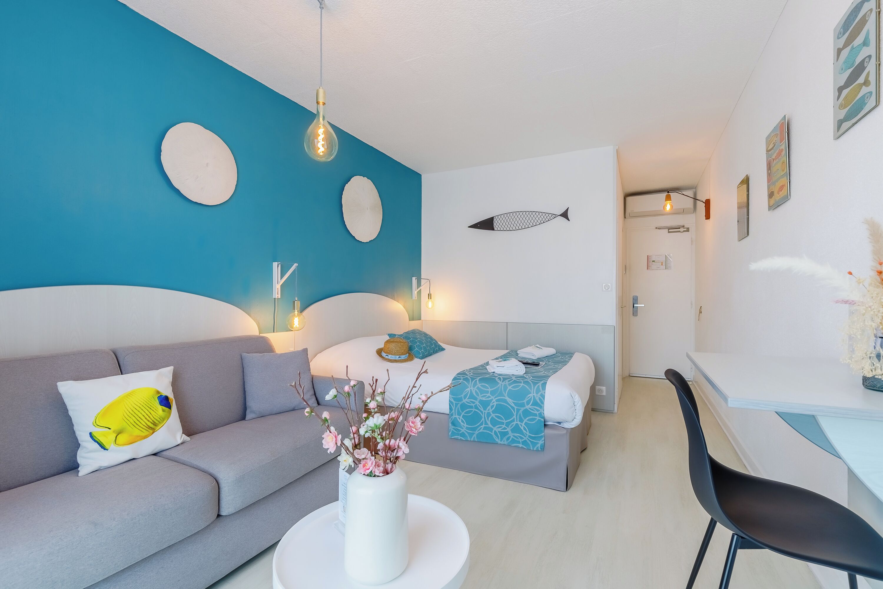 Une chambre d’hôtel moderne avec un mur d’accent bleu, un lit recouvert d’une courtepointe à motifs bleus et blancs, un canapé gris avec un coussin jaune, un vase blanc contenant des fleurs et une chaise noire.