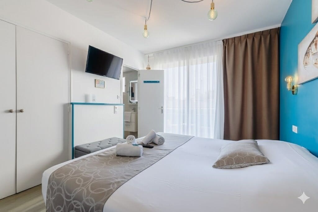 A hotel room with a bed, a TV mounted on the wall, a desk with a chair, and a window with curtains.