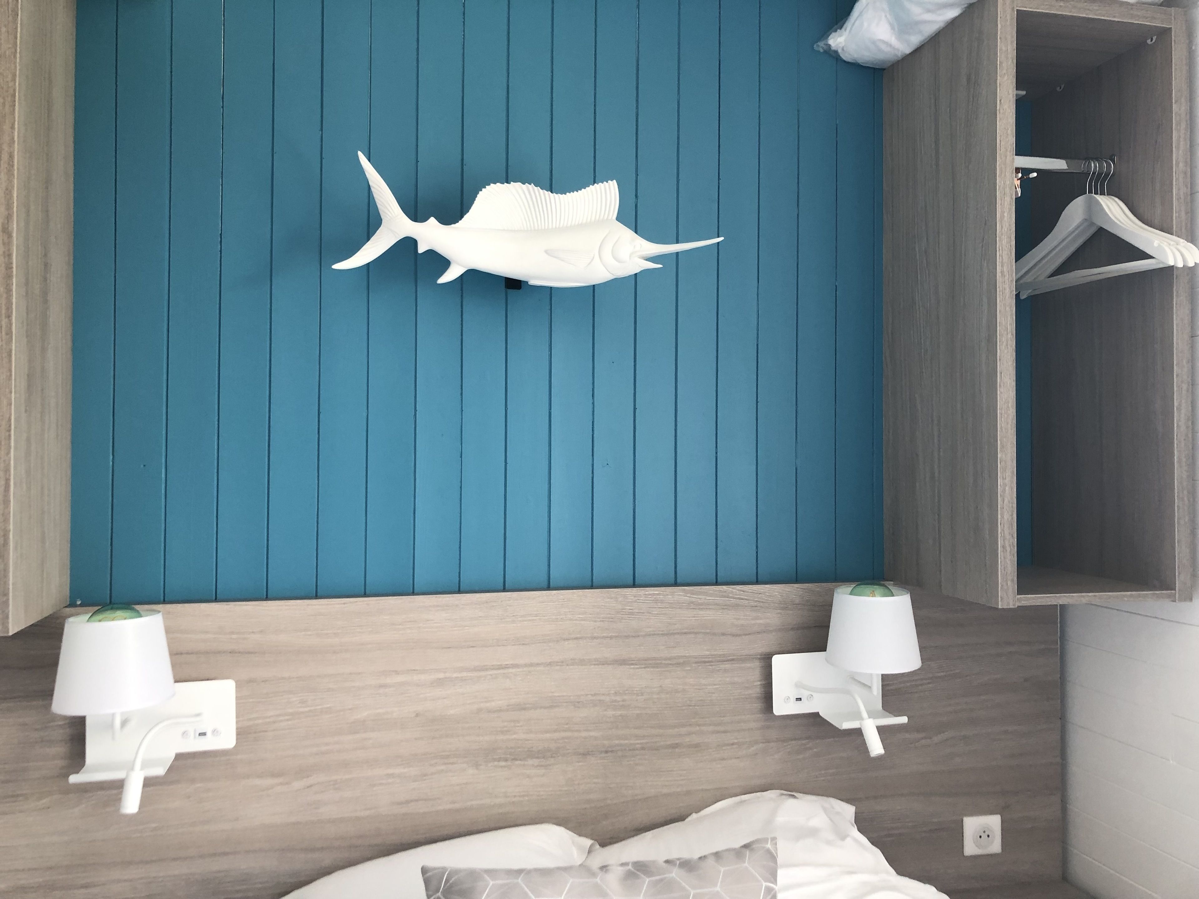 Quarto com parede azul, cabeceira de madeira, duas mesas de cabeceira com luminárias, cama com colcha estampada em branco e cinza, e prateleira branca fixada na parede com decoração de peixe branco.