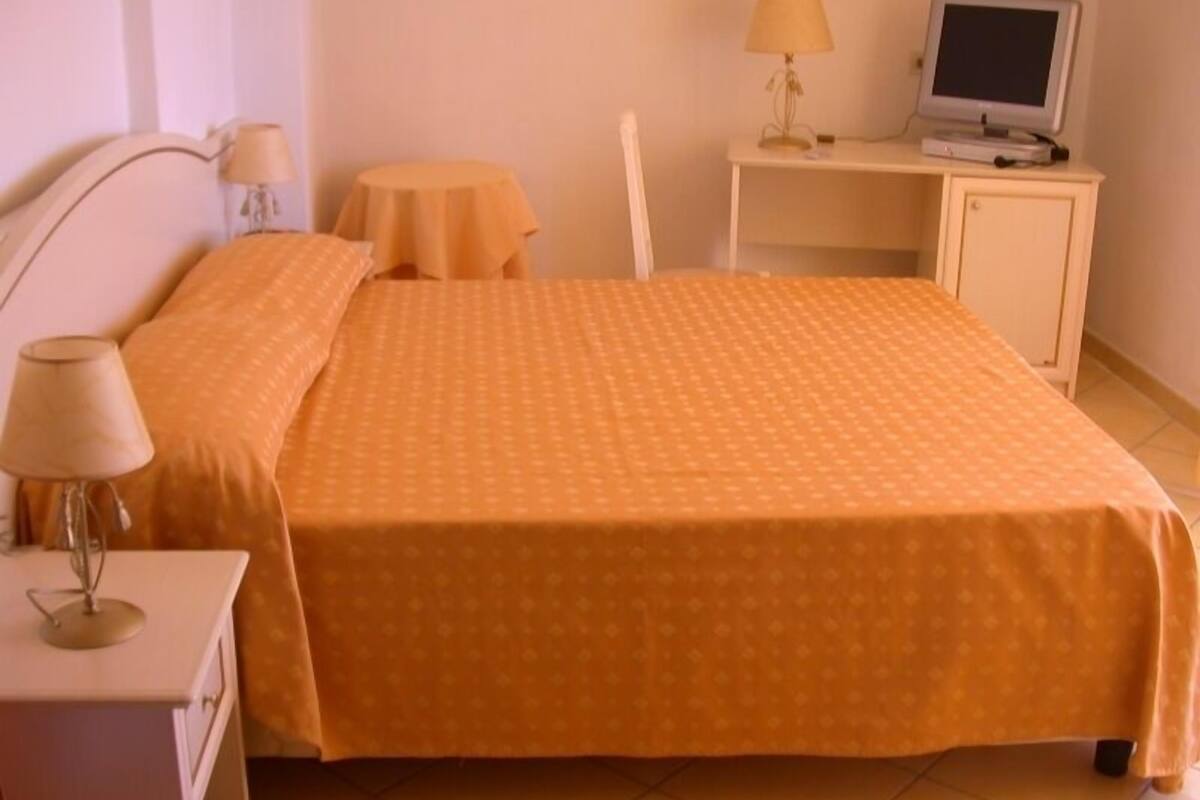 Una cama bien hecha con colcha naranja, una mesita de noche con lámpara, una pequeña mesa redonda con lámpara, y un escritorio con monitor de computadora.