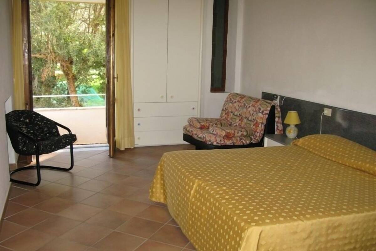 Un dormitorio con cama, una silla, armario y ventana con cortinas.