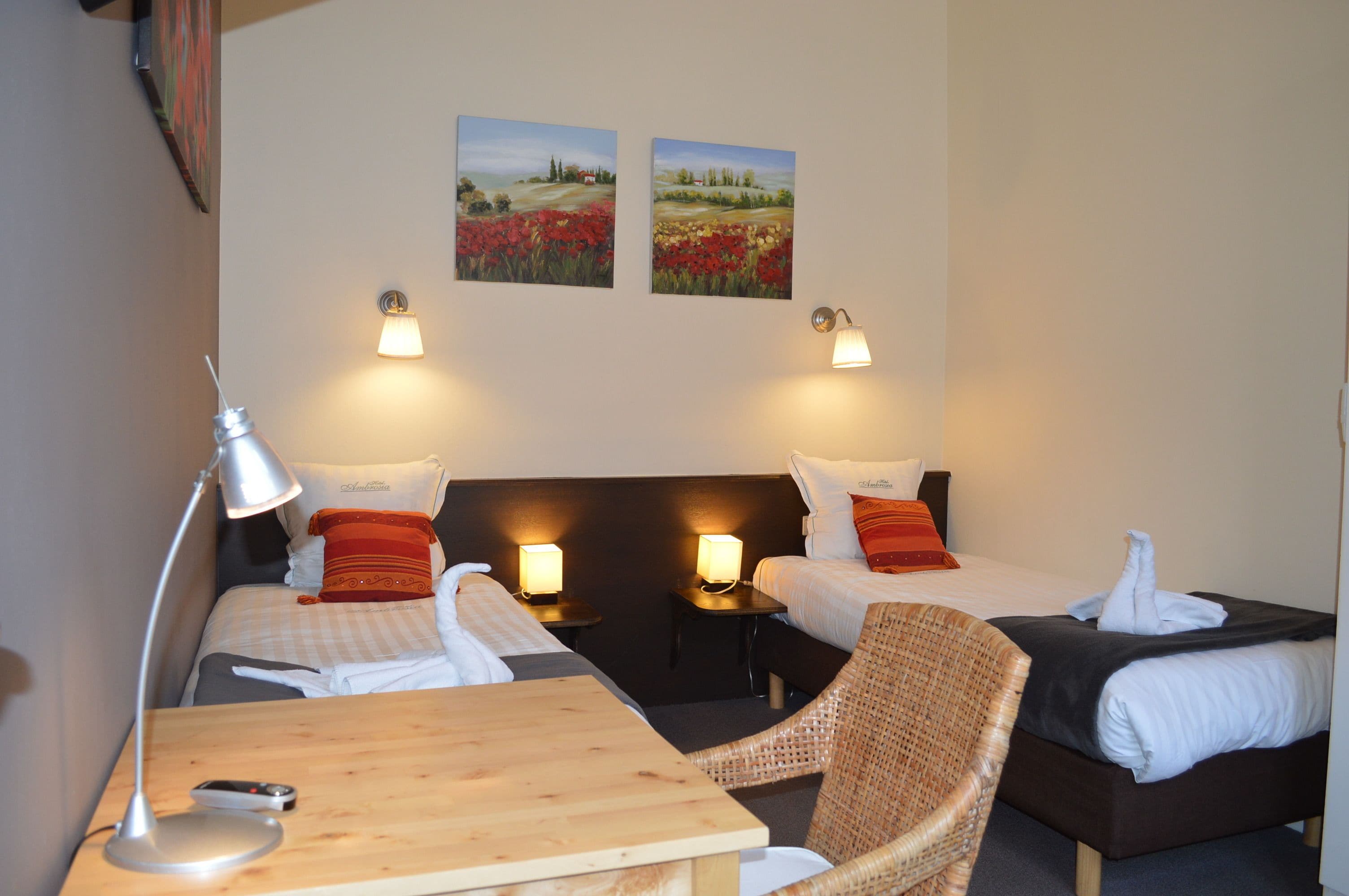 A hotel room with two beds, a wooden table, a wicker chair, and two wall-mounted lamps.