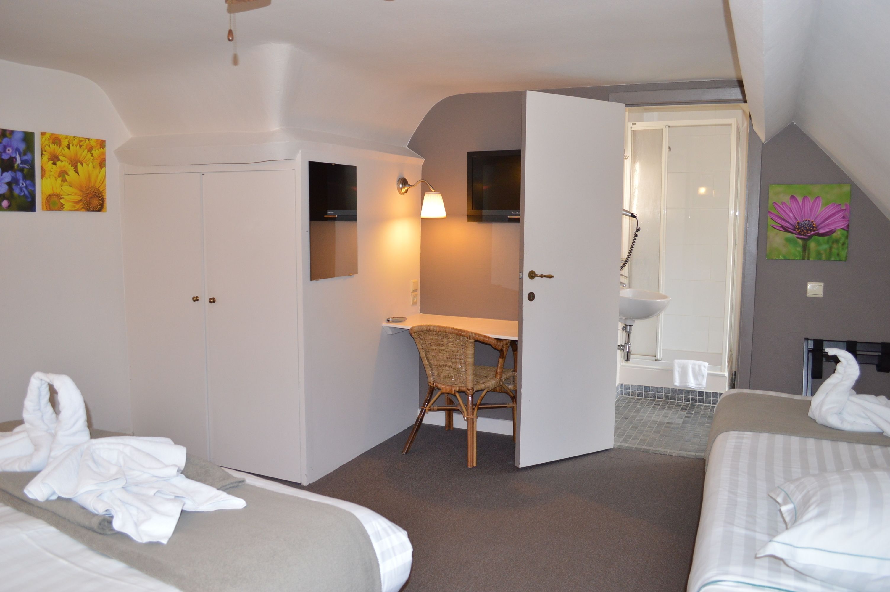 A hotel room with two beds, a desk, a chair, a wardrobe, a bathroom with a sink and mirror, and artwork on the walls.