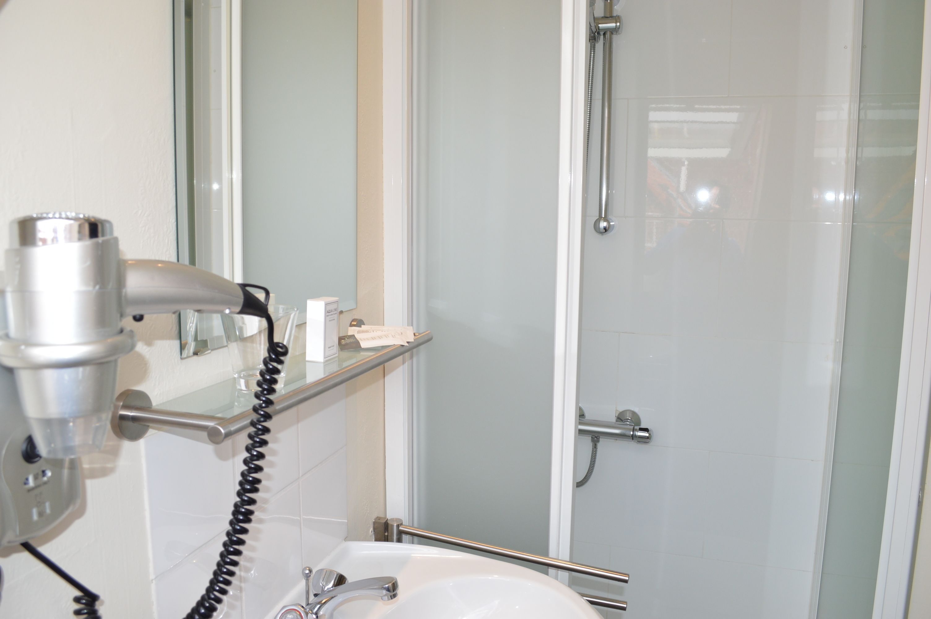 Un baño con ducha, lavabo, secador de pelo y repisa de vidrio con artículos de aseo.