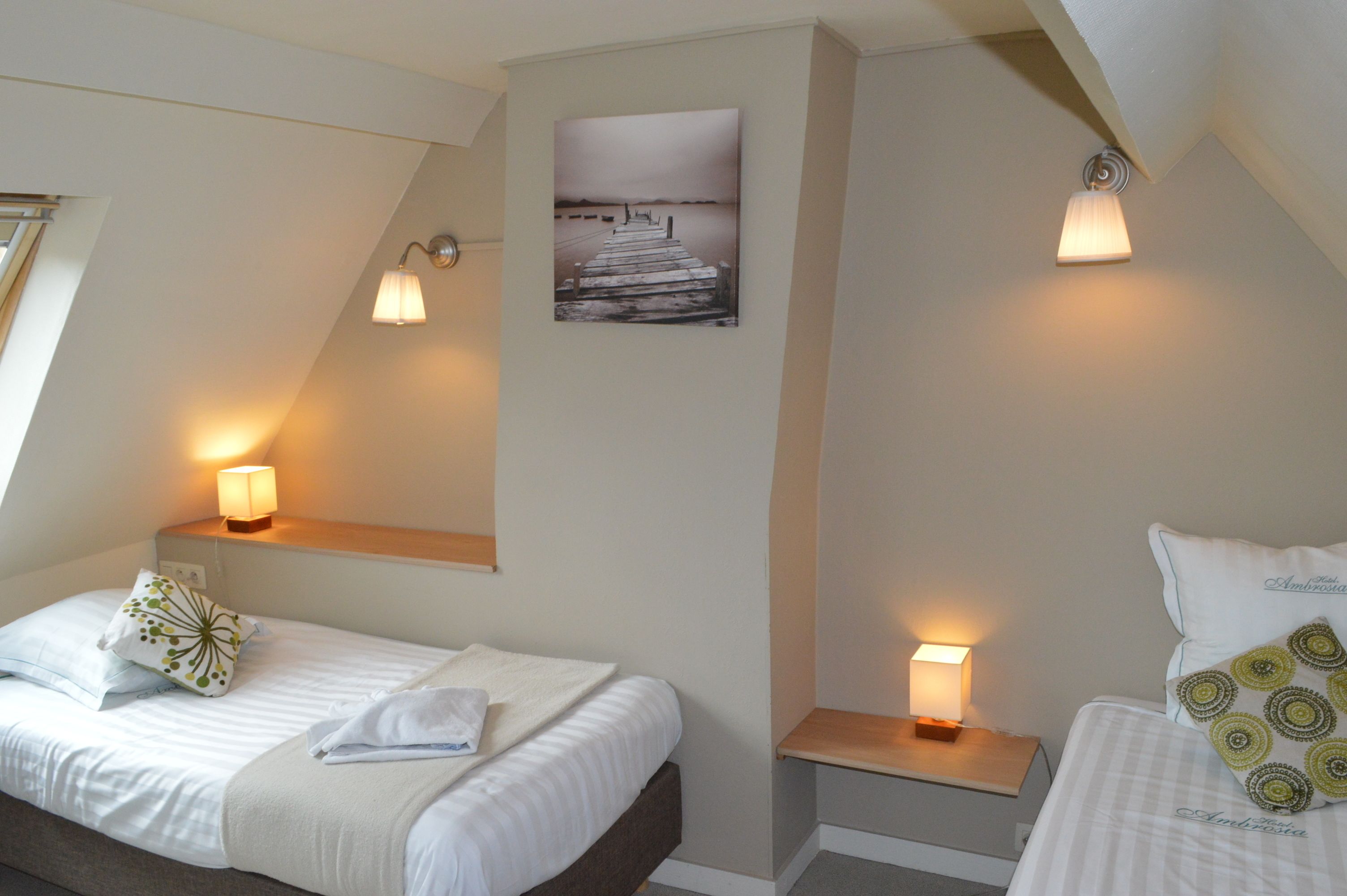 Habitación de hotel con dos camas, cada una con una mesita de noche y lámpara, un cuadro en la pared y una ventana que proporciona luz natural.