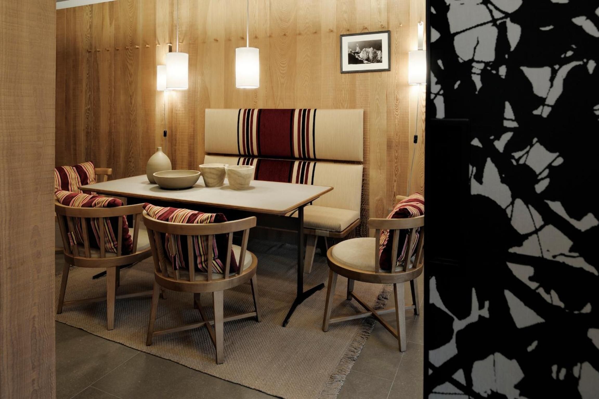 Un coin repas avec un mur en bois, un banc et quatre chaises dotées de coussins à rayures. Deux lampes suspendues sont placées au-dessus de la table.