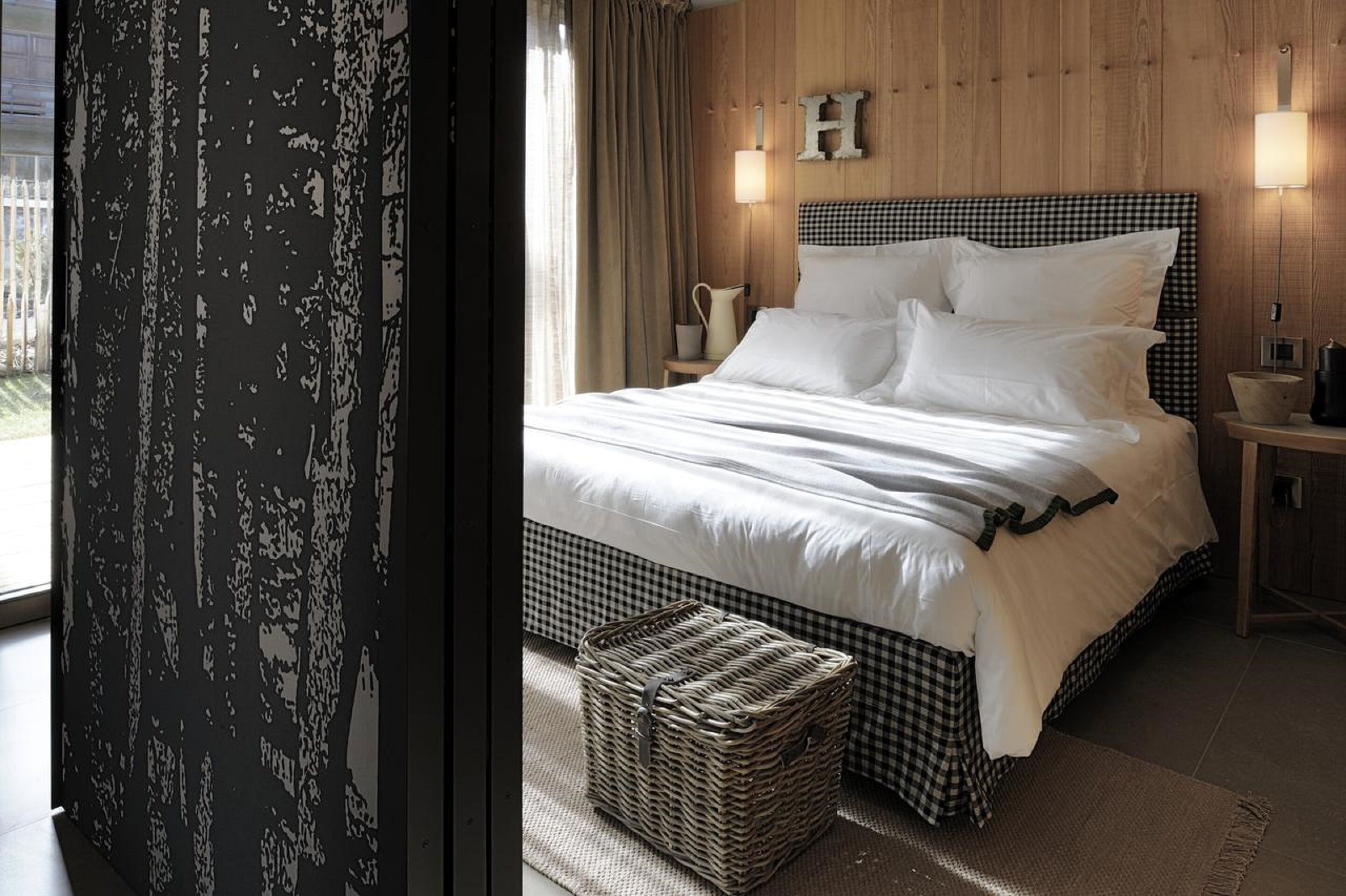 A bedroom with a checkered bedspread, a bedside lamp, a wicker basket, and a wooden headboard with a letter 'H'.