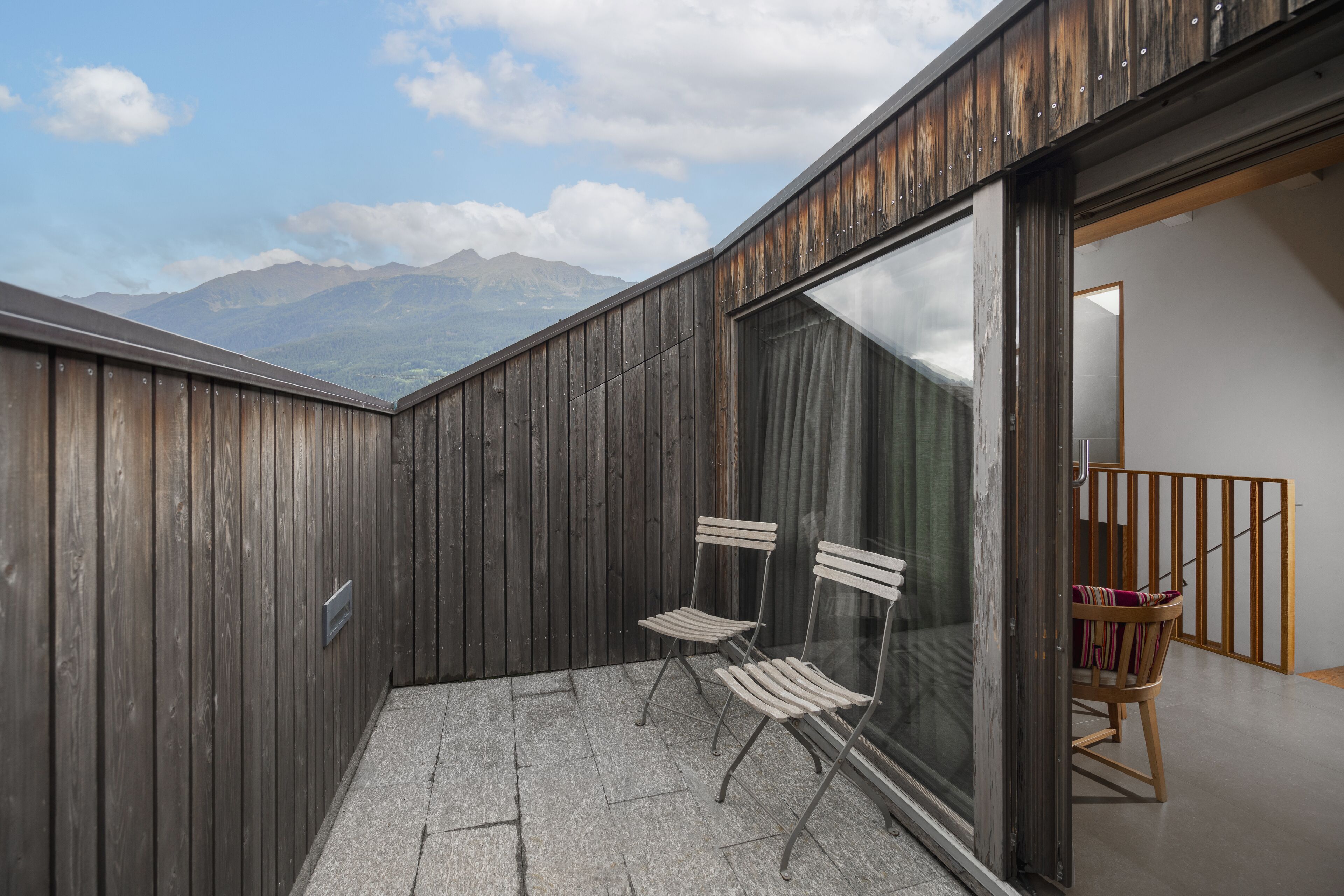 A balcony with wooden walls, two metal chairs, and a view of mountains.