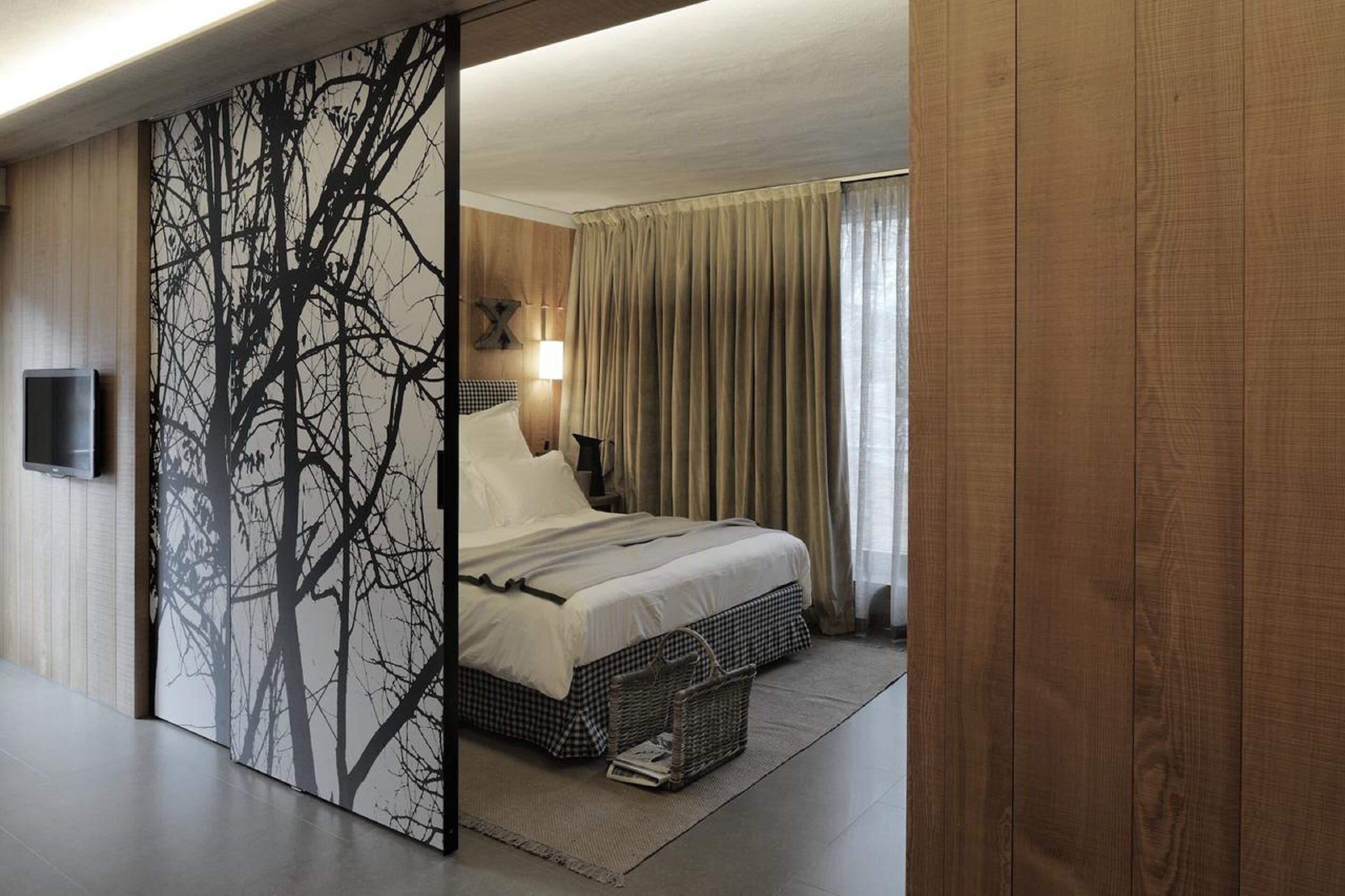 Una camera d'albergo moderna con un letto grande, una televisione a parete e vista all'esterno tramite una finestra con tende.