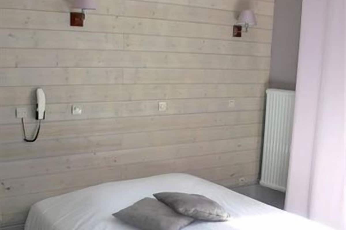 A bedroom with a wooden wall, a white bed, two pillows, a bedside lamp, a phone, and a radiator.