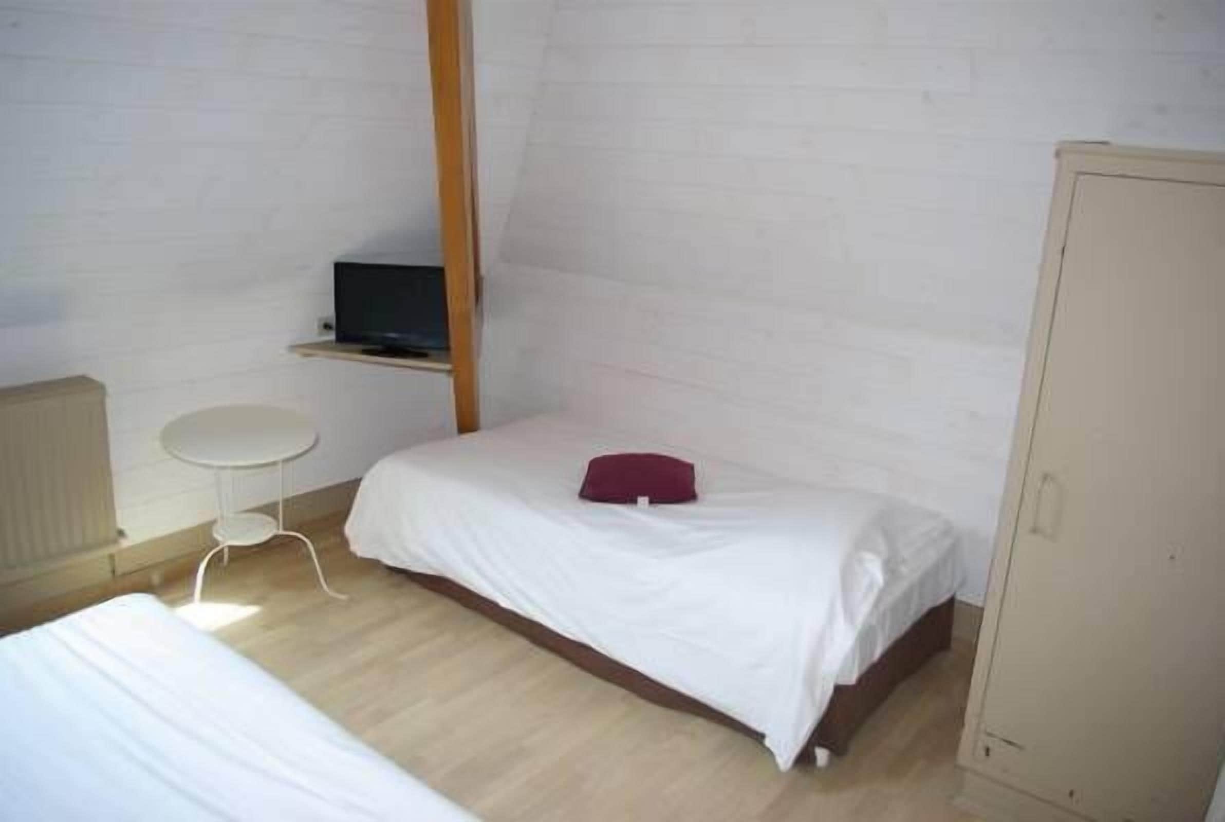Een kleine eenpersoonskamer met een wit bed, een klein tafeltje, een televisie op een plank en een kledingkast.