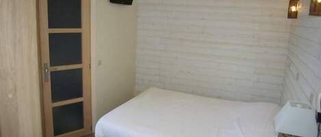 Un dormitorio con una cama individual, una puerta de madera, un televisor montado en la pared y una mesita de noche con lámpara.