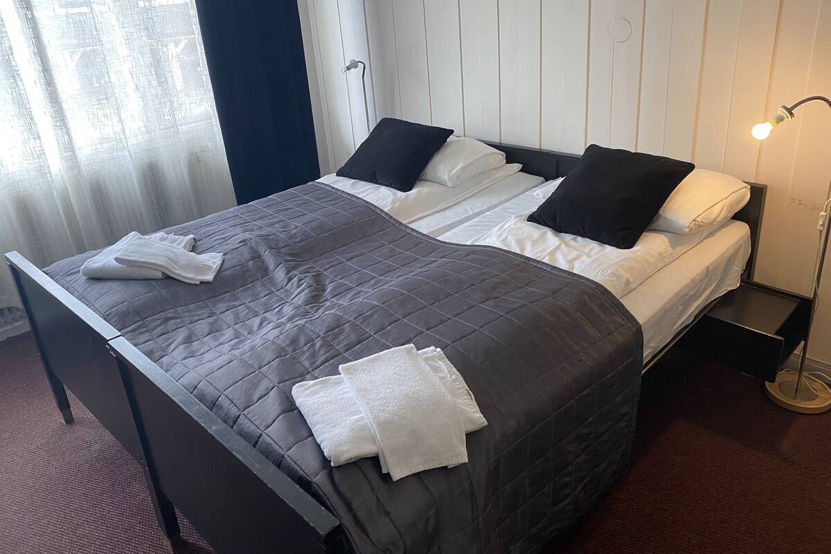 A neatly made bed with a dark quilt and white linens, a bedside table with a lamp, and a window with sheer curtains.