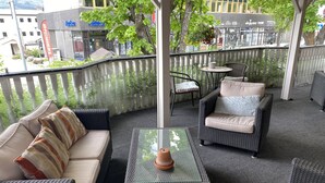 En balkon med fletmøbler, heriblandt en sofa og stole, et lille bord med en vase, og udsigt over bybilledet.