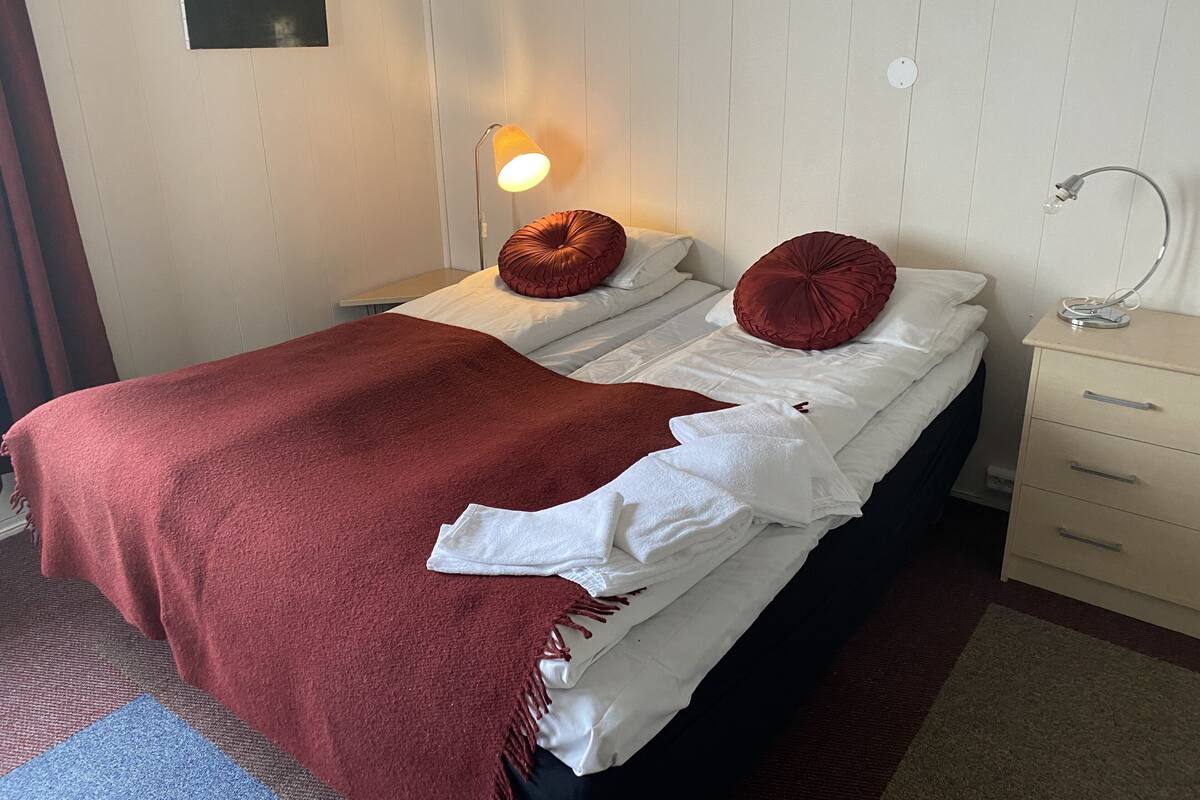 A neatly made bed with a red blanket, two red pillows, and a white towel. There is a bedside lamp on the nightstand.