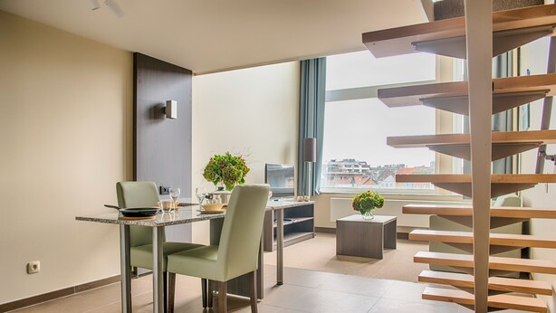 A modern living room with a dining area, a staircase, and a large window offering a city view. - Arass Hotel & Business Flats (Antwerp)