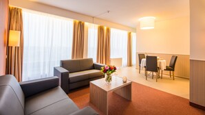 A modern hotel room with a sofa, a coffee table with a vase of flowers, a dining area with a table and chairs, and large windows with curtains. - Arass Hotel & Business Flats (Antwerp)