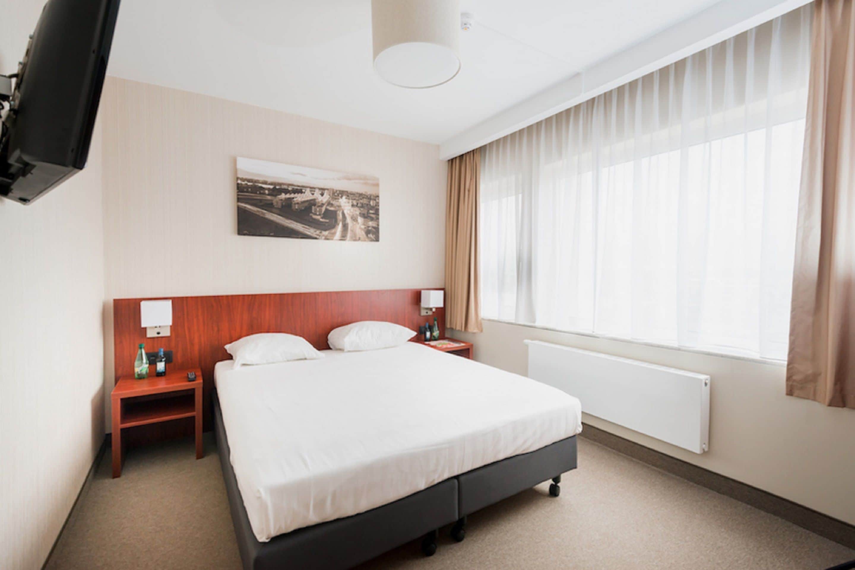 Habitación de hotel con una cama grande, un televisor de pantalla plana en la pared, una mesita de noche con lámpara y una ventana con cortinas traslúcidas.