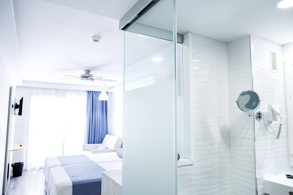 A modern hotel room with a glass shower enclosure, a bed with a blue accent, and a ceiling fan.