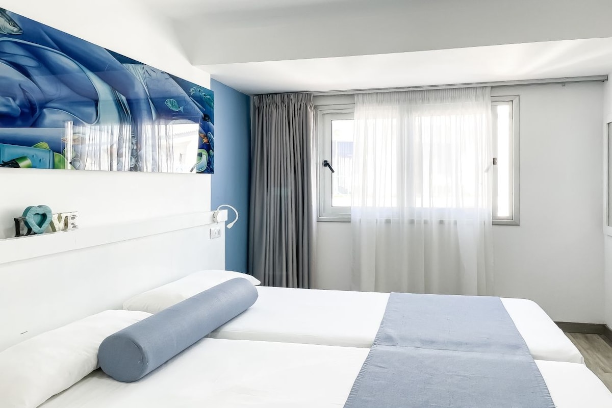 Una habitación de hotel moderna con cama, un ventanal con cortinas, una pared con un mural azul y una mesita de noche con lámpara.