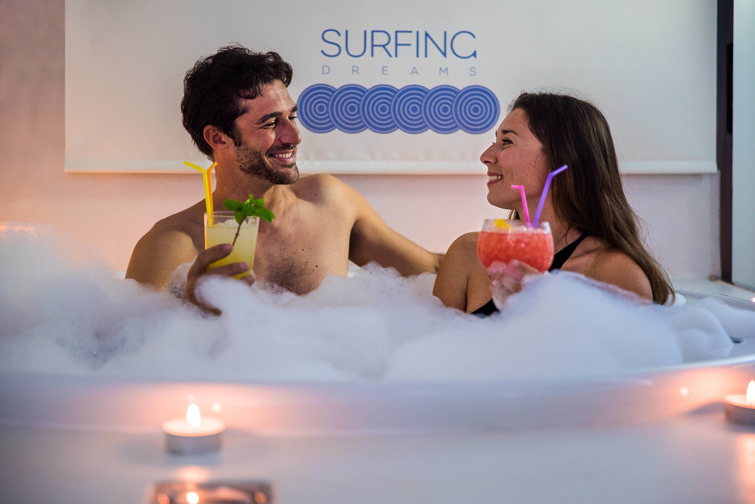 Two people in a hot tub with drinks, surrounded by candles.