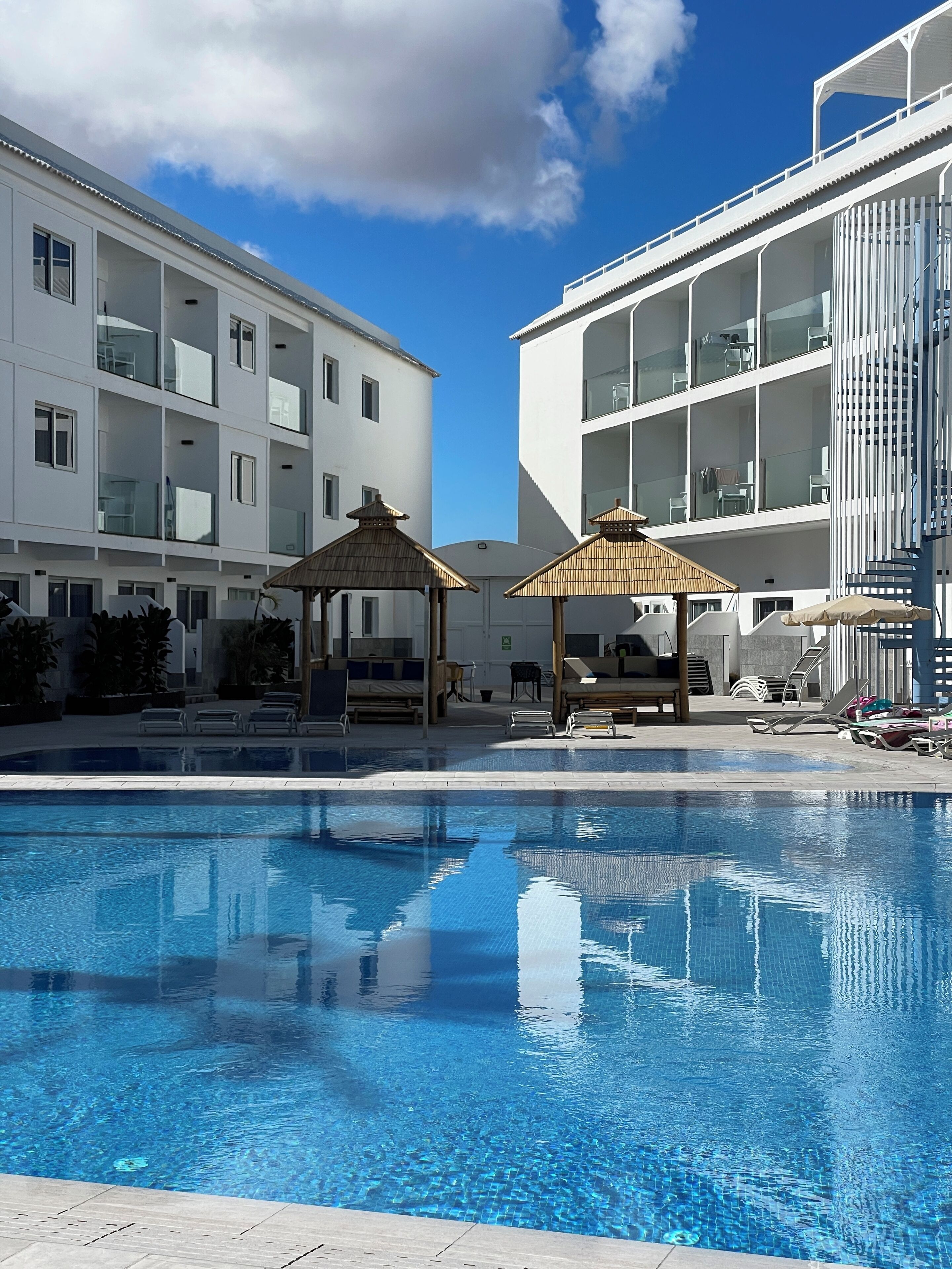 Una piscina con sillas de descanso y sombrillas, rodeada de edificios modernos con balcones.