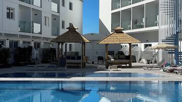 Una piscina con sillas de descanso y sombrillas, rodeada de edificios modernos con balcones.