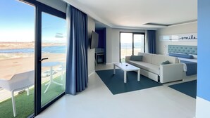 A modern hotel room with a large sliding glass door to a balcony, a white sofa, a small table, and a bed with a blue headboard. - Corralejo Surfing Colors Hotel&Apartments (La Oliva)