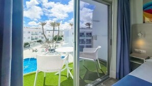 A hotel room with a sliding glass door leading to a balcony with a pool and palm trees. - Corralejo Surfing Colors Hotel&Apartments (La Oliva)