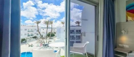 Habitación de hotel con puerta corredera de vidrio que da a un balcón con piscina y palmeras.