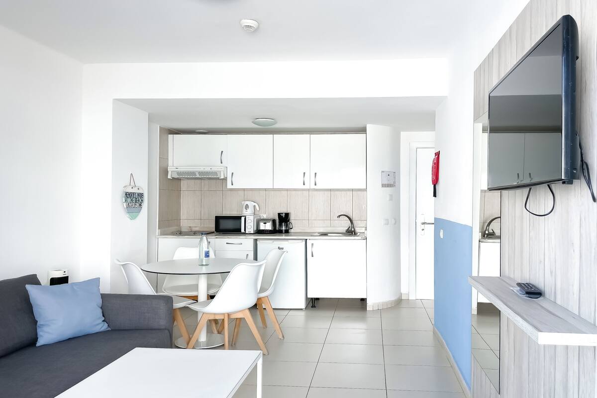 A modern kitchen with white cabinets, a microwave, and a coffee maker. There is a dining area with a table and chairs, and a sofa with cushions. A flat-screen TV is mounted on the wall.