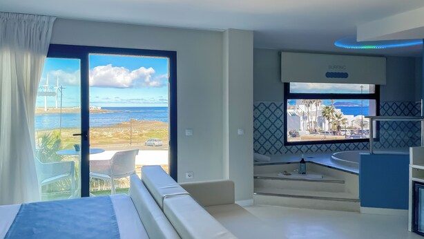 A modern hotel room with a large bed, a balcony with a view of the sea and wind turbines, a small kitchen area, and a TV mounted on the wall. - Corralejo Surfing Colors Hotel&Apartments (La Oliva)