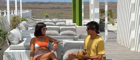 Un espacio de estar al aire libre techado con mobiliario blanco, una mesa con una bebida y vistas a un paisaje árido.