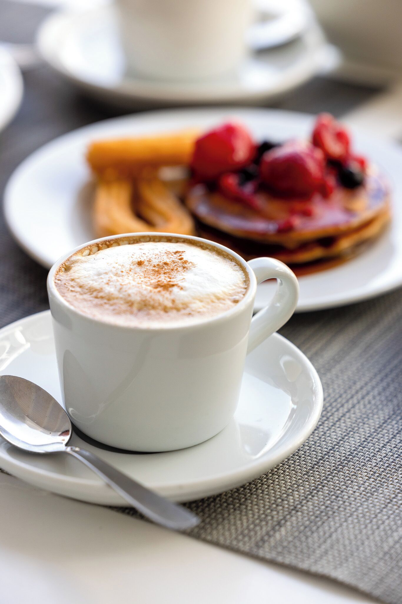 Una taza blanca de café con una cuchara sobre un plato, un plato con tortitas y bayas, y una taza desenfocada al fondo.