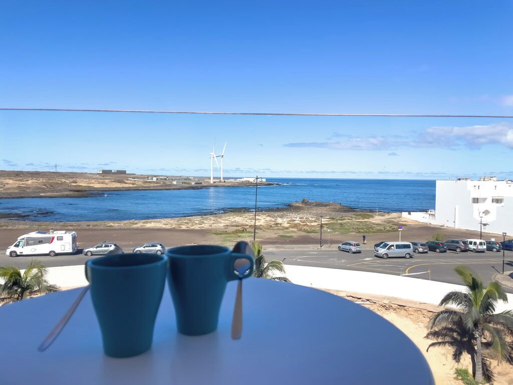 A balcony with two blue cups, overlooking a coastal road with parked cars and wind turbines in the distance.