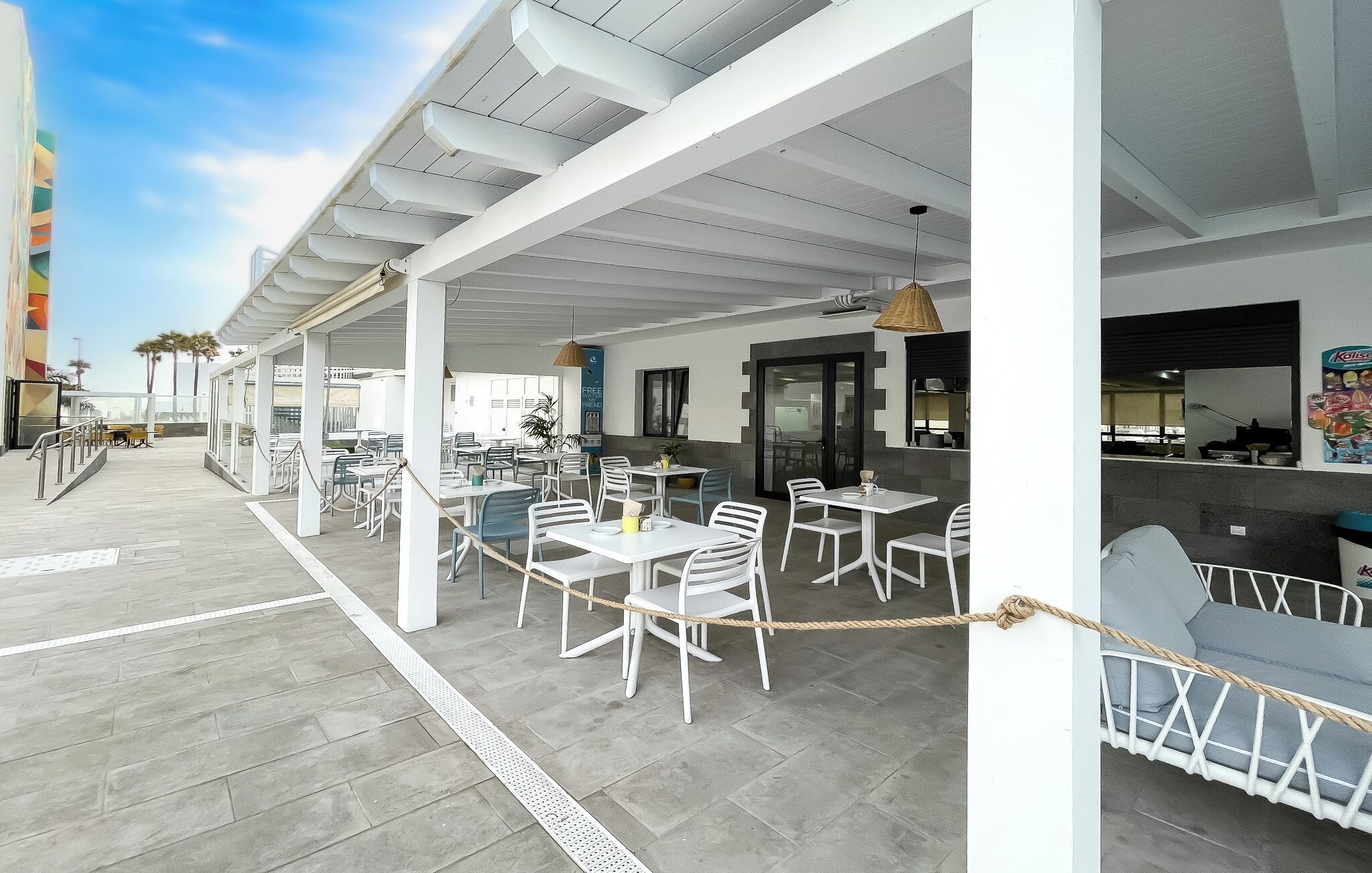 An outdoor cafe with white tables and chairs, a rope barrier, and a view of the ocean.