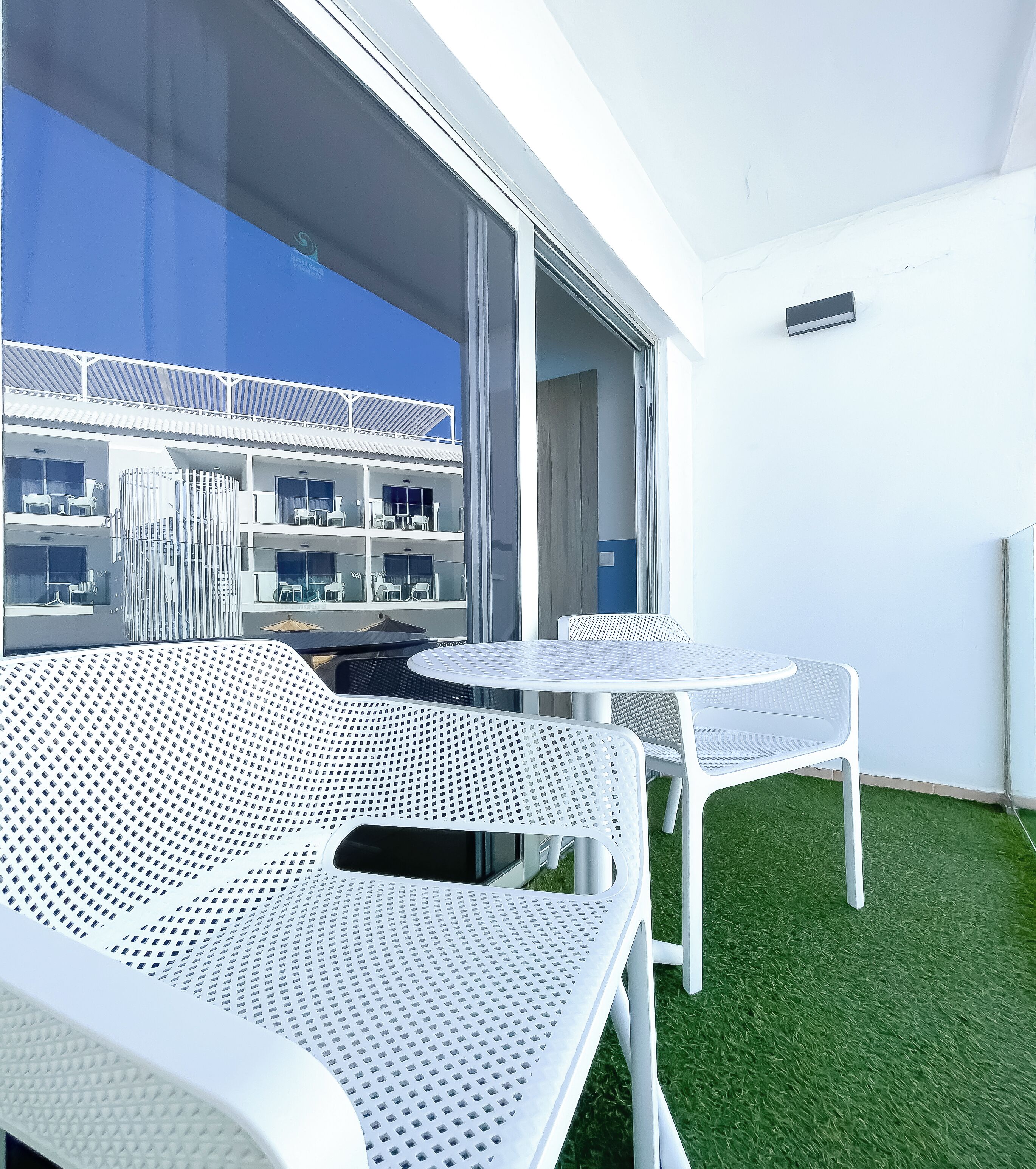 A balcony with white metal chairs and a table, a glass door leading to an interior space, and a view of a building with balconies.