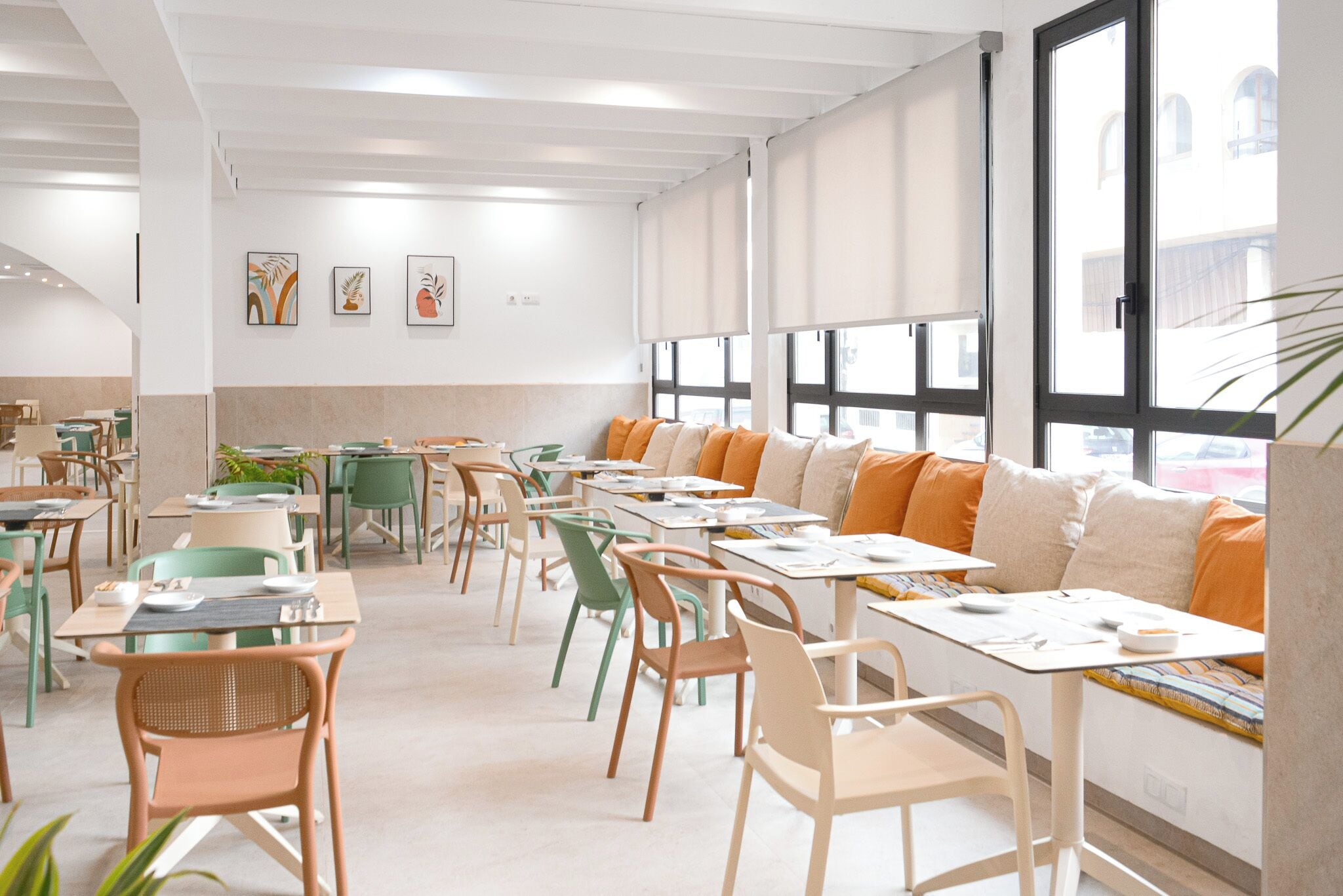 A spacious dining area with multiple tables and chairs, large windows with blinds, and artwork on the walls.