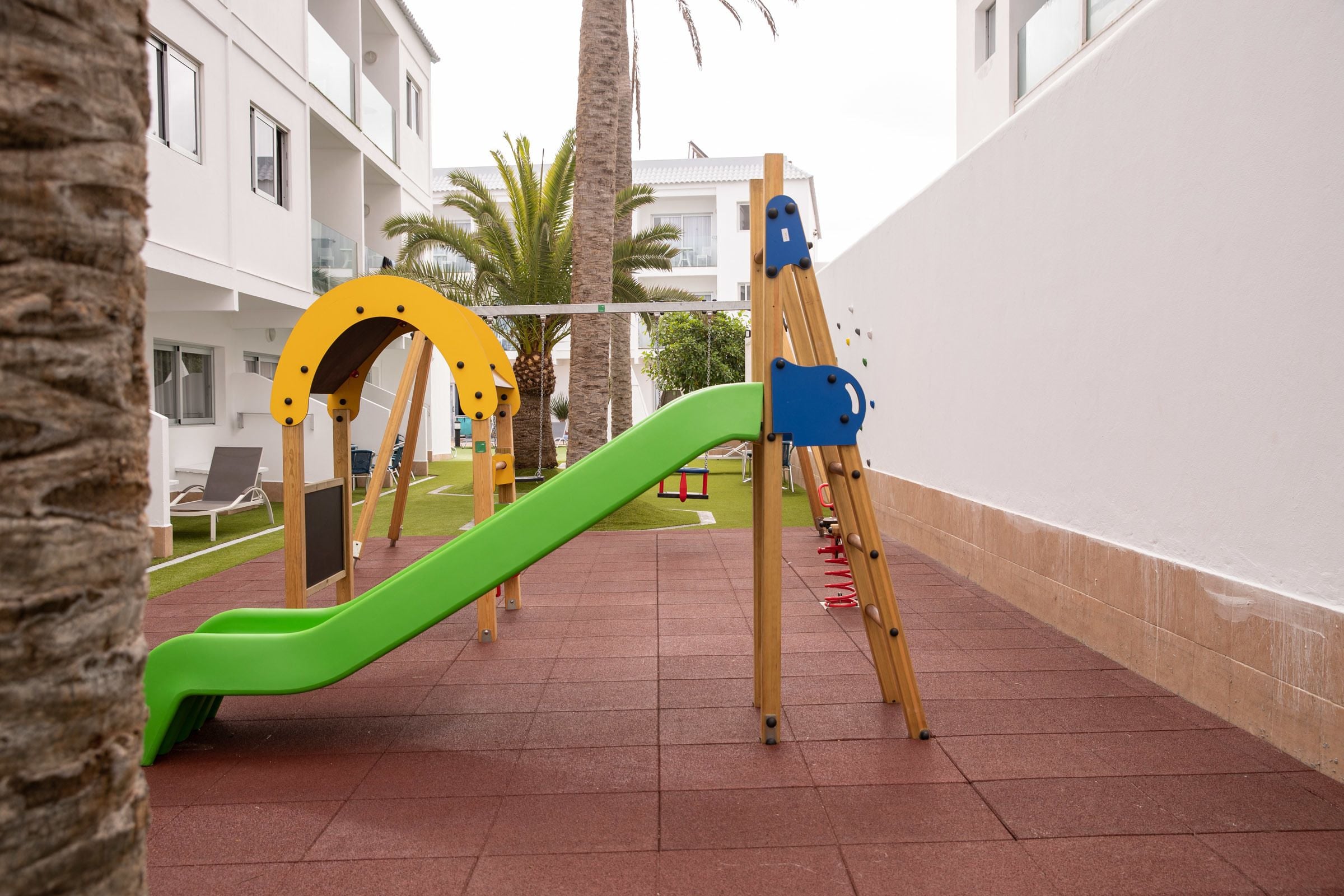 A playground with a green slide and climbing structures, surrounded by a building with balconies and palm trees.