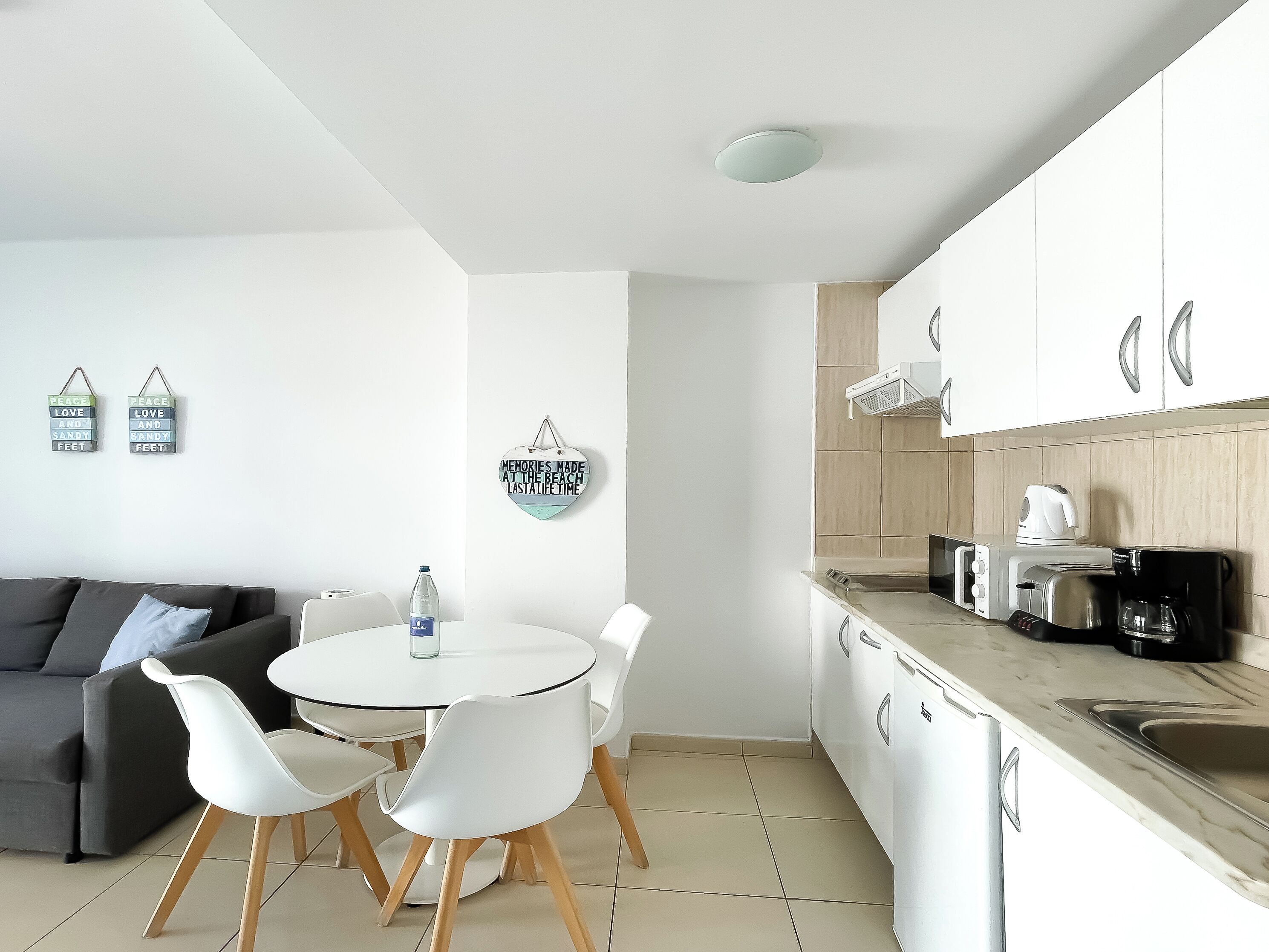 A modern kitchen with white cabinets, a round dining table with white chairs, and a grey sofa in the living area.