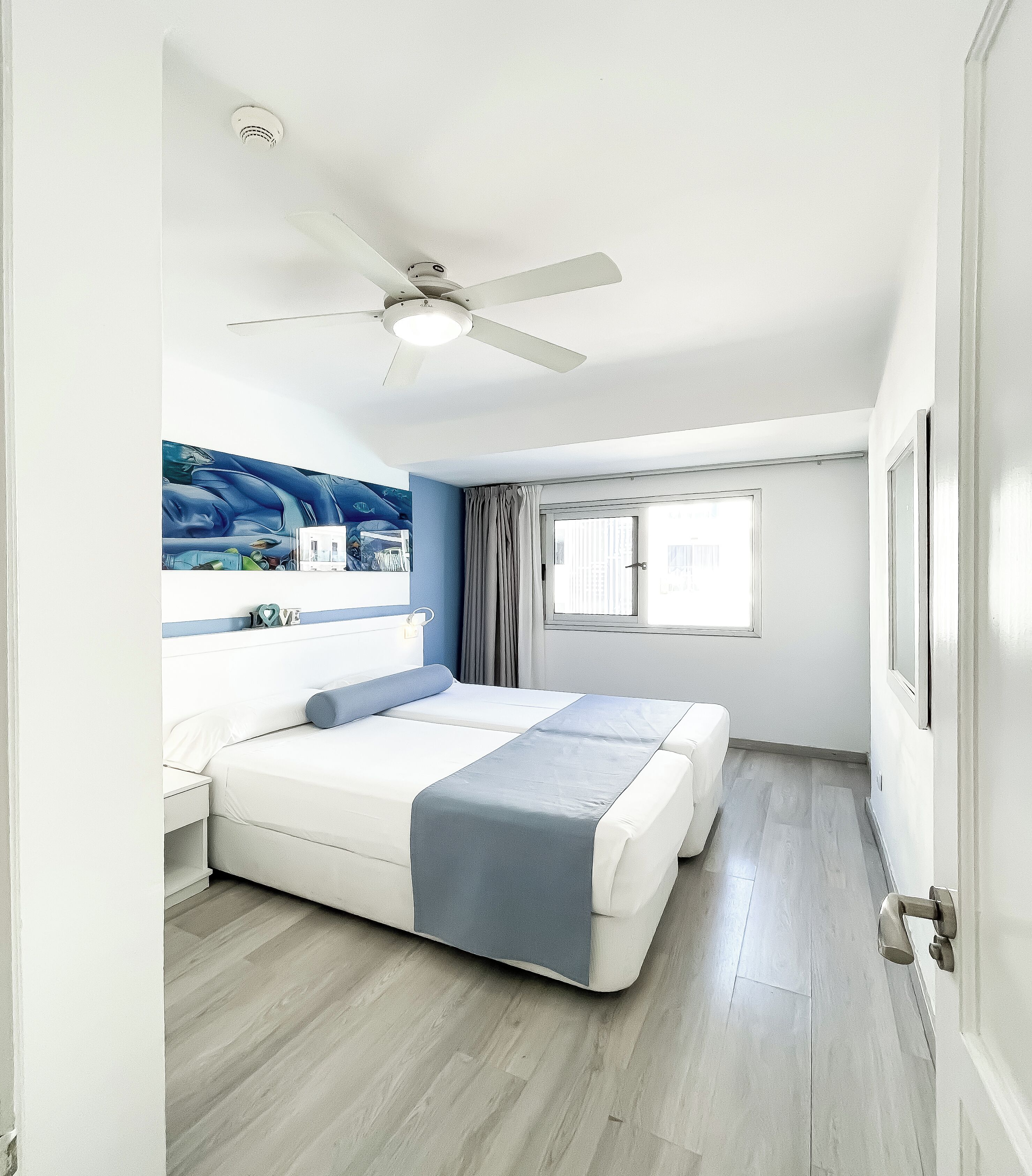 A bedroom with a bed, a ceiling fan, a window, and a view of the city.